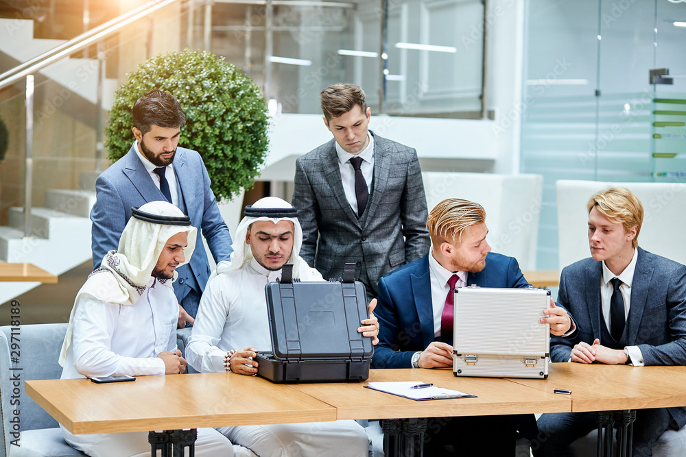 Caucasians in suits evaluate money by sheikhs, sheikhs in white suits look at modern technology, opening black case. Business concept Caucasians in suits evaluate money by sheikhs, sheikhs in white suits look at modern technology, opening black case. Business concept