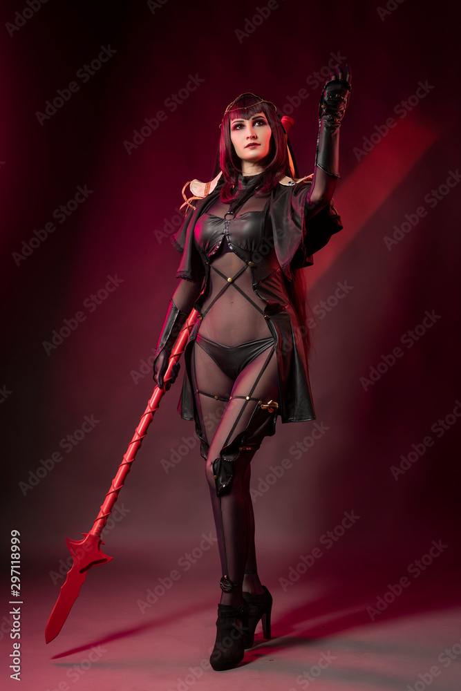 Beautiful leggy busty cosplay girl wearing an erotic leather costume posing with a fake spear on a red background in theatrical smoke.