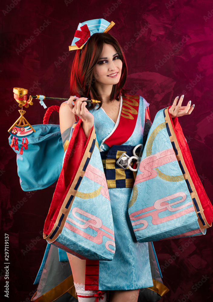 Naklejka premium Beautiful leggy busty cosplayer girl wearing a stylized Japanese kimono costume cheerfully posing holding a fake pipe on a blue background.