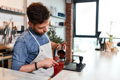 Strict busy barman stands at caunter in apron, puts ground coffee into tamper, lookins aside, expressis attentiveness, working hard concept photo