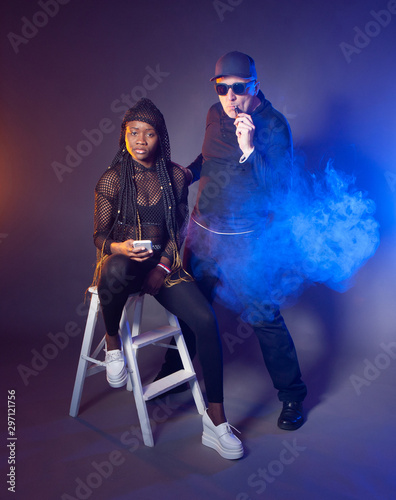 Black and white concept. Dark-skinned girl in black clothes sitting on a white stool. A man with white skin smokes VAPE. Communication of different people. African girl with a white phone.