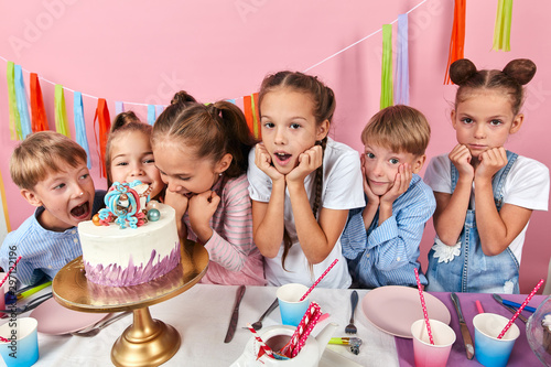 children wth fists under their chind standing around the cake waiting for their portion, isolated pink background, studio shot