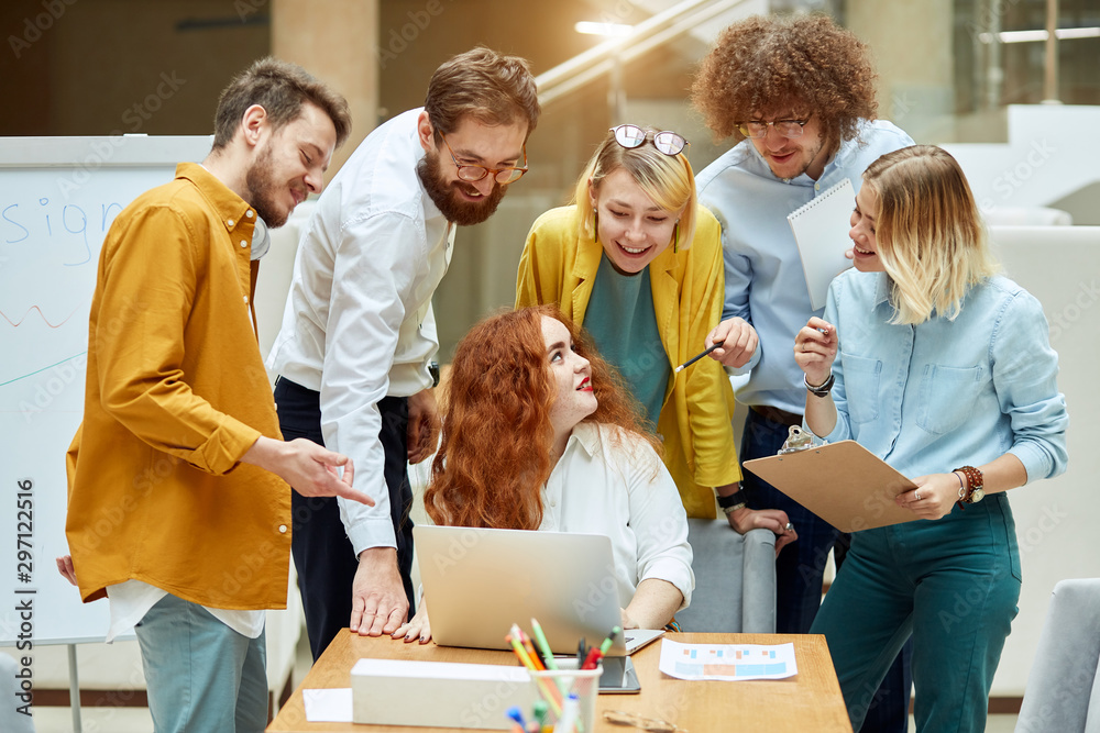 Charming red haired woman sits at table with computer on it, turns hands back, looks smilingly at optimistic group of designers, asks for advice, in modern brightly lighted office