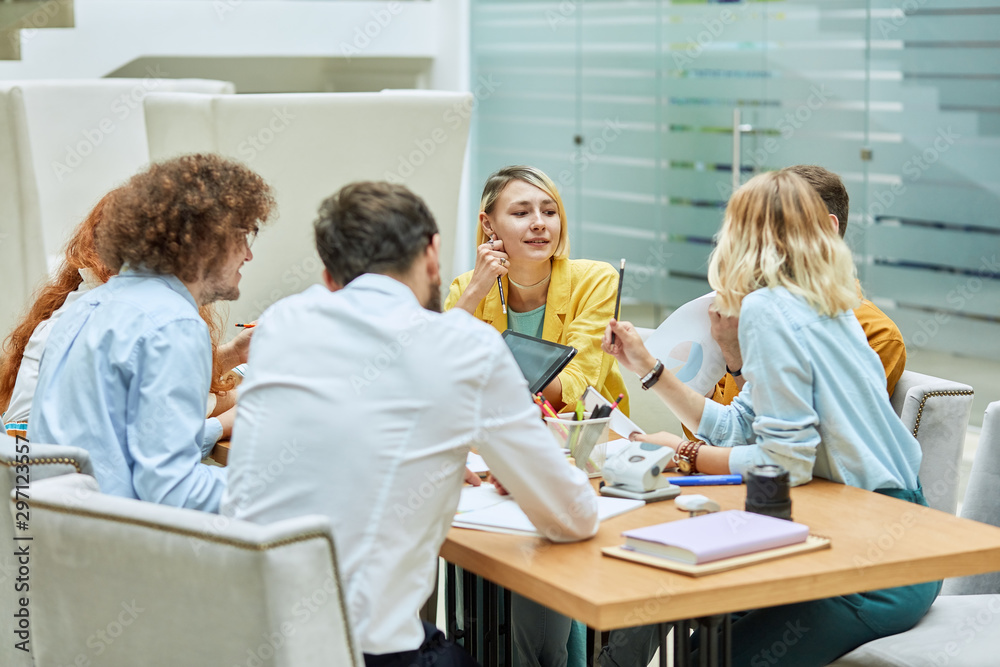 Hard working team of bright designers brainstorming in modern office, pretty blonde woman sits at table, dressed in colourful clothres, holds pencil, keeps hand on gentle cheek, looks away