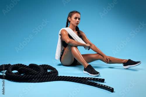 Beautiful fit pesive thoughtful young girl with white towel on her shoulder sitting on a floor. battle ropes in the background of the photo. free time, spare time. lifestyle photo