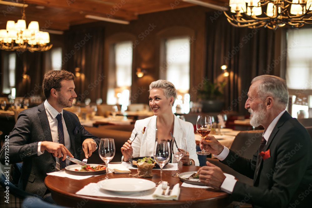 Good joyful friends sit at round wooden table in cozy soft chairs in bright lighted restaurant, drink alcohol, eat tasty food, smile happily, posing over large windows with thick curtains