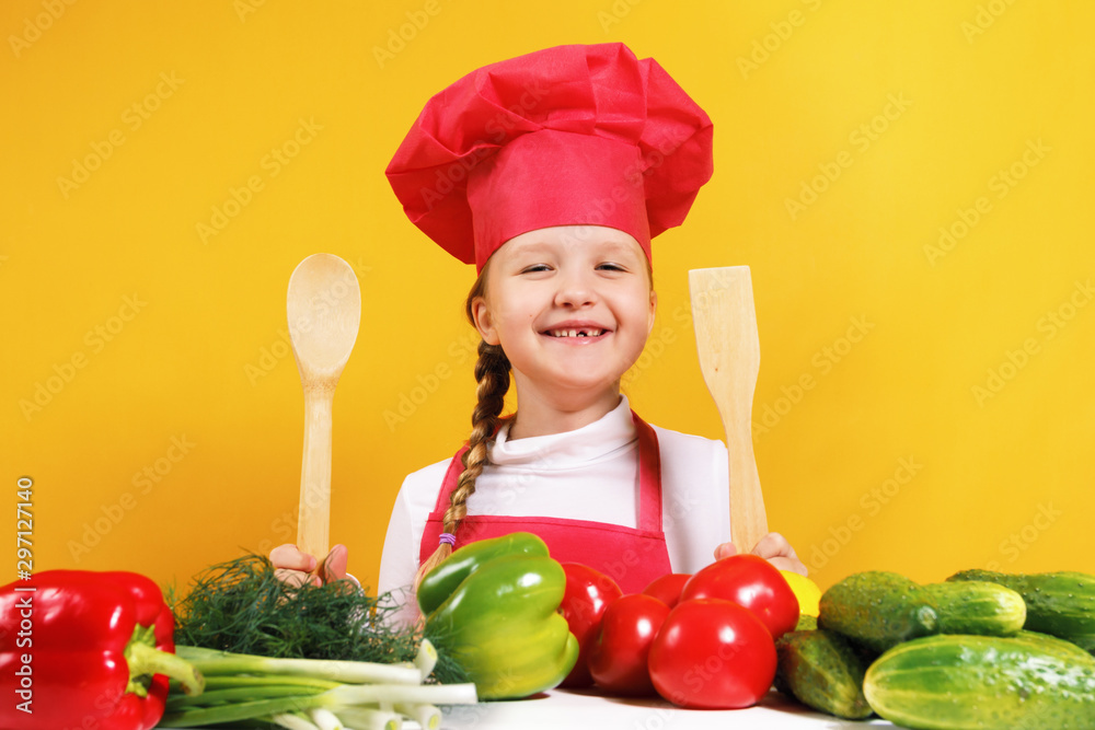Beautiful little girl chef on a yellow background. The child sits at a table with vegetables and holds in his hands a wooden spoon and a spatula Beautiful little girl chef on a yellow background. The child sits at a table with vegetables and holds in his hands a wooden spoon and a spatula