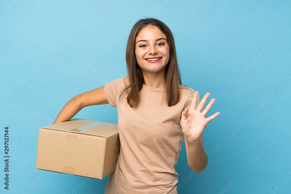 Young brunette girl over isolated blue background holding a box to move it to another site and saluting Young brunette girl over isolated blue background holding a box to move it to another site and saluting