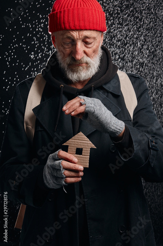 Obraz na plátne Senior street man wearing coat and red hat, holding house made by cardboard dream about own shelter