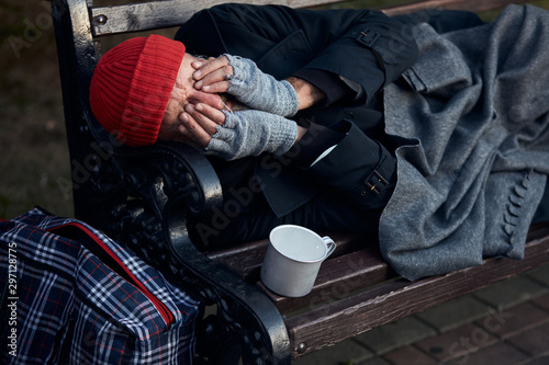 Penniless senior man lying on park bench, trembling from the cold, closed face with hands. Cup for collecting money, coins next to him.