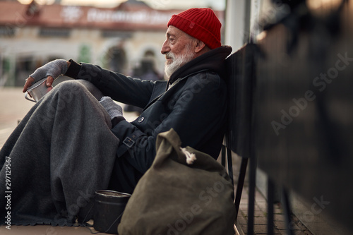 Side view on beggar male on street begging, waiting help by citizens. Senior homeless man in old clothes sit on floor in cold weather photo