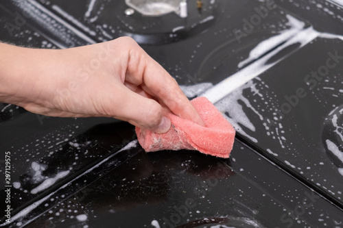 Female hand washes gas stove, glass ceramic surface. Red sponge and white foam.