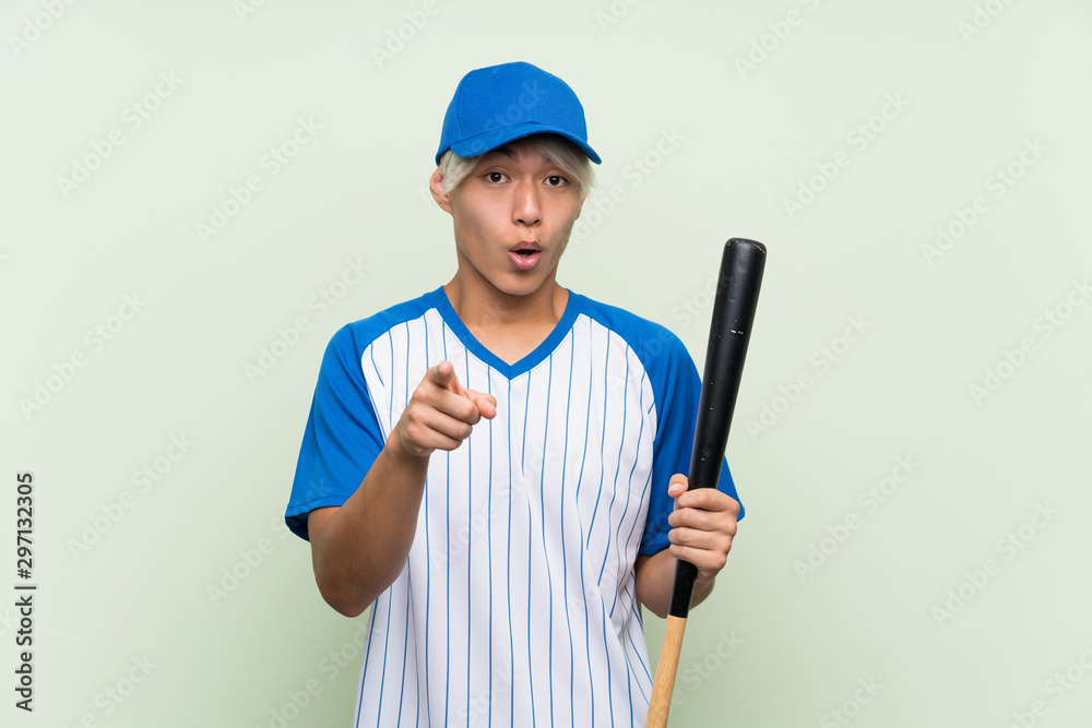 Young asian man playing baseball over isolated green background surprised and pointing front Young asian man playing baseball over isolated green background surprised and pointing front