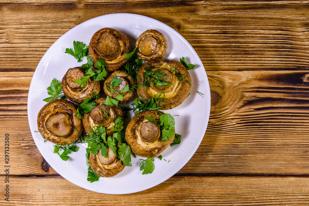 Plate with baked champignons, dill and parsley on a wooden table. Top view Plate with baked champignons, dill and parsley on a wooden table. Top view