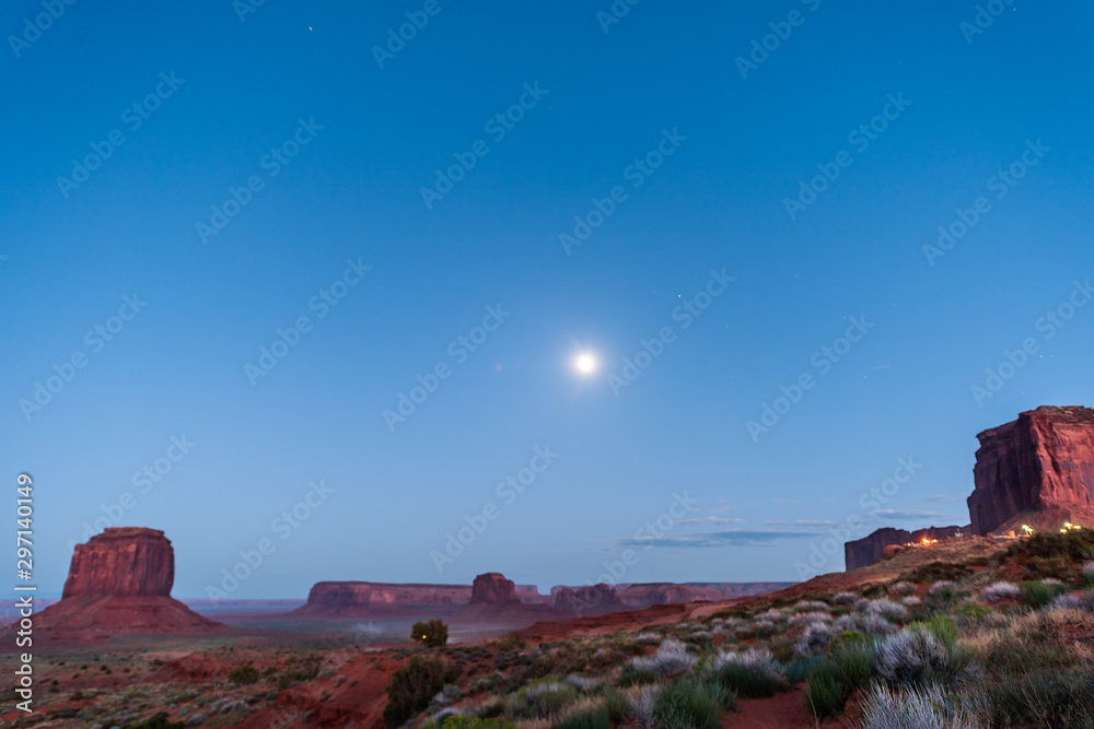 Butte formations wide angle view in Monument Valley during blue twilight dark night in Arizona with cars on dust dirt road and moon in sky