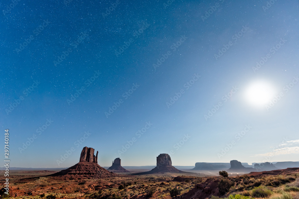 Famous buttes wide angle view in Monument Valley during blue twilight dark night with stars in Arizona dust road and bright moon in sky