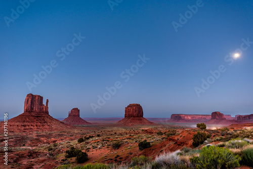 Famous mesa butte formations with red rock color in Monument Valley canyons during twilight dark night in Arizona with cars on dirt road and moon in sky