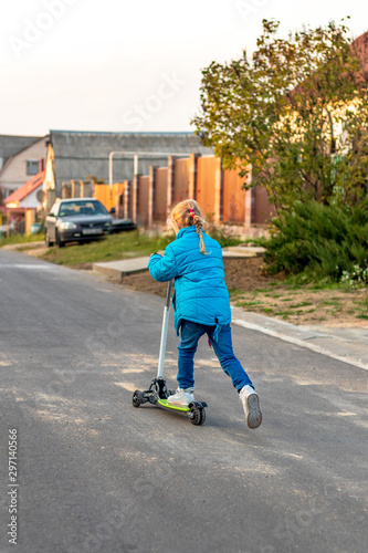 back view. girl in a blue jacket riding a scooter. blond hair color. she rolls her foot on the board.