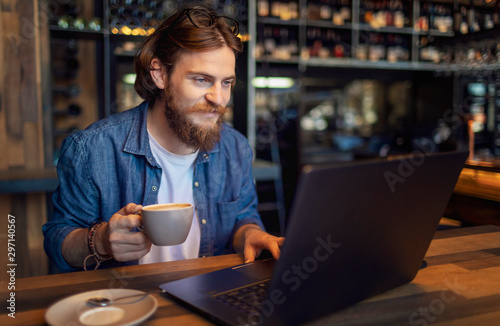 Young bearded freelancer checking email via laptop while having morning coffee in a loft bar
