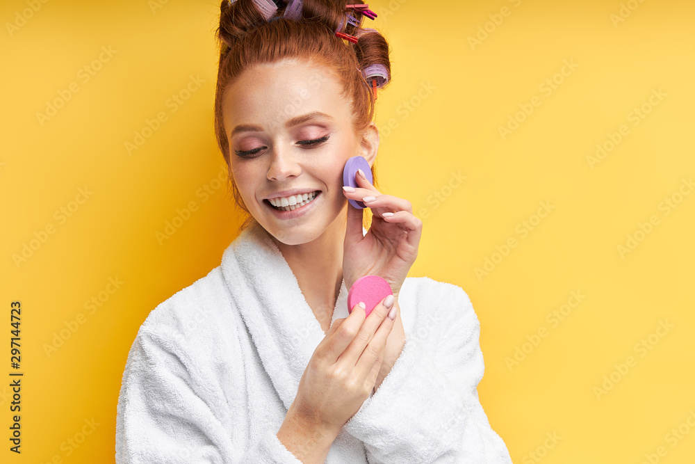 Careful beautiful woman isolated over yellow background, holding beauty sponges near face. Attractive redhead woman in bathrobe. with curlers - obrazy, fototapety, plakaty 