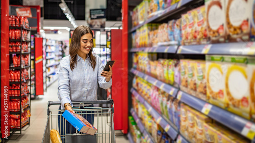 Pretty young woman buying groceries in a supermarket/mall/grocery store. Shopping list. Smiling beautiful woman stands with phone near products in store photo