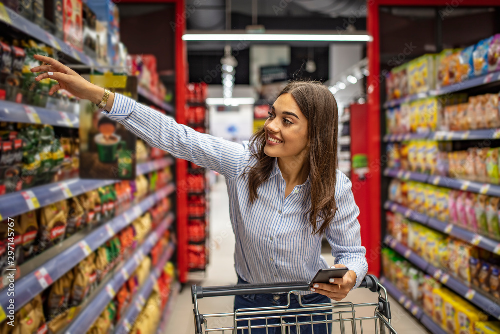 Young cheerful woman in supermarket with shopping trolley choosing products and using phone. Looking aside. The housewife chooses food for dinner at home on the background of shelves in a supermarket.