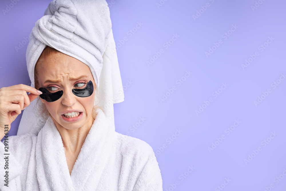 Caucasian female in bathrobe and towel removing patches from eye. Cosmetology and skin care concept. Caucasian female in bathrobe and towel removing patches from eye. Cosmetology and skin care concept.