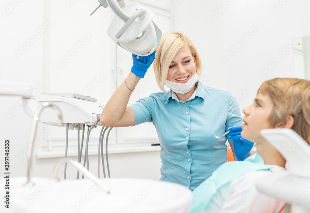 Smiling blonde female dentist adjusting dental reflector lamp before examining a boy