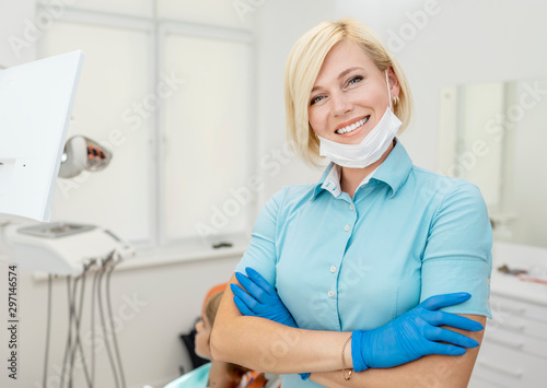 Portrait of cute blonde dentist in blue scrubs and gloves posing in her workplace
