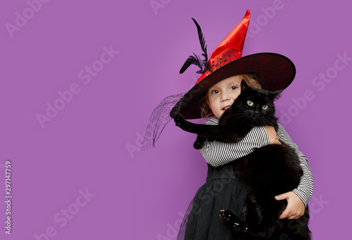 Child in Halloween costume. Kids trick or treat. Little girl holds a black cat on purple, violet background. Baby in witch hat and kitten. Autumn season holiday decoration. 