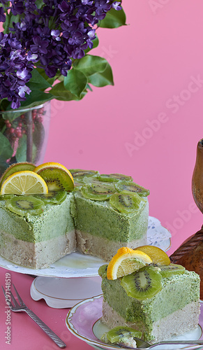  Front view of a piece of cake with kiwis and cashews. Accompanied by the rest of the cake, violet flowers and a pink background.