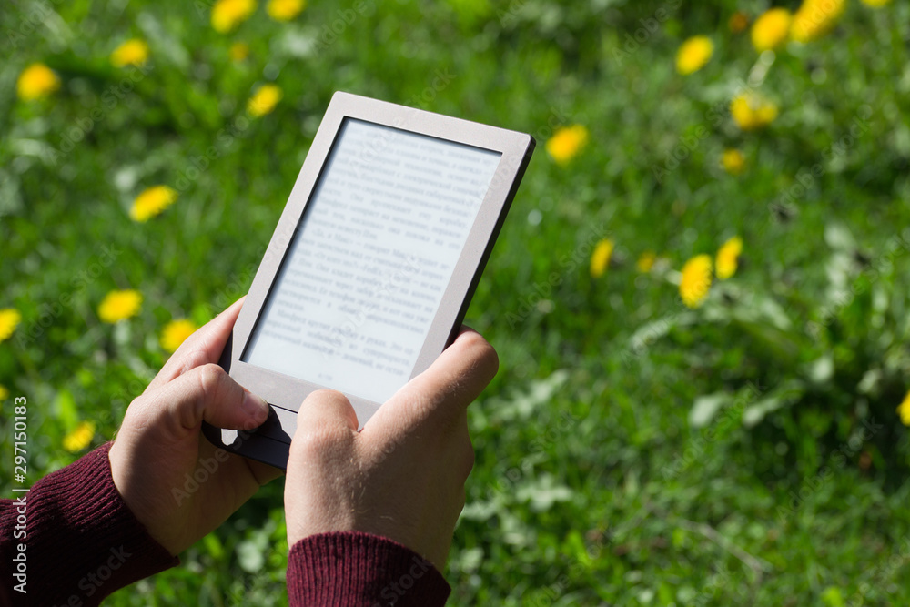 A man is reading an electronic book at spring sunny day. Blurred text. Only his hands are on the picture. Dandelion field at background.