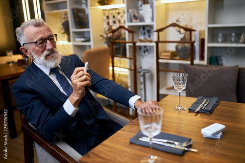 Serious grey-haired elderly man in tuxedo smoke IQOS in restaurant, explore modern way smoking cigarette