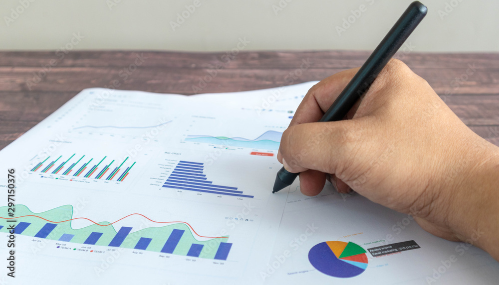 A man's hand holding a pen is about to write something on the line graph, circle graph, and various color chart graphs printed on a white paper. Placed on a wooden patterned work desk A man's hand holding a pen is about to write something on the line graph, circle graph, and various color chart graphs printed on a white paper. Placed on a wooden patterned work desk