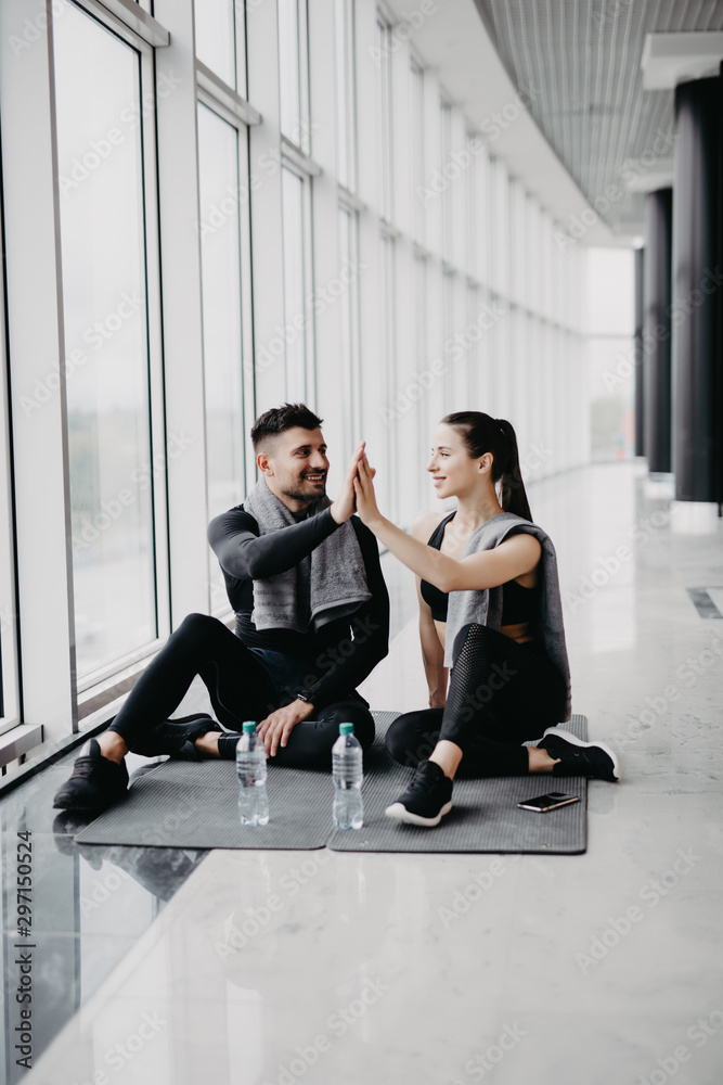 Fit young couple in sportswear smiling and high fiving together while sitting on a gym floor after working out