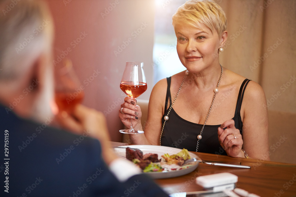 Caucasian senior woman in black dress fine dinner food with man in elegant restaurant. Female raised a glass and going to drink. Rear view on husband in tuxedo.