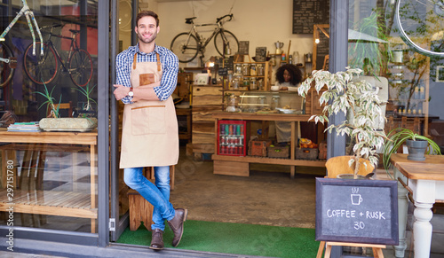 Canvas Print Smiling young barista leaning against the door of a cafe