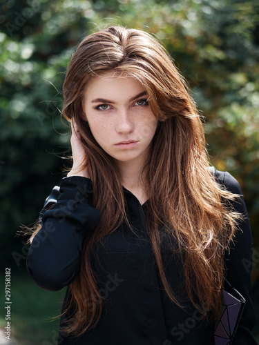 Closeup portrait of young natural beautiful redhead woman in black blouse touching her long natural hair against blurred green foliage park background windy weather