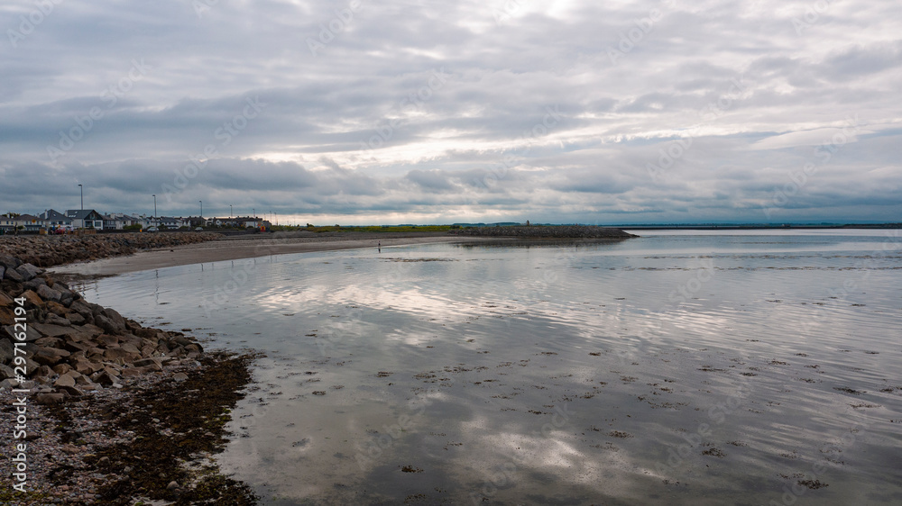 View of Salthill beach and Mutton island on a cloudy day, with rocks in the foreground and mountains in the background.