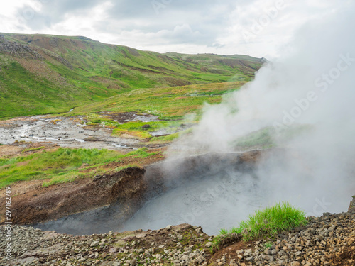 Reykjadalur valley with hot springs river and pool with lush green grass meadow and hills with geothermal steam. South Iceland near Hveragerdi city. Summer sunny morning  blue sky.