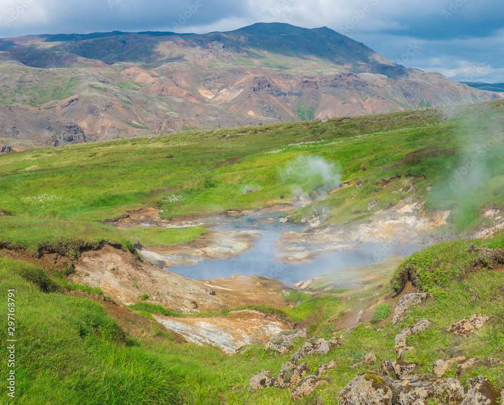 Reykjadalur valley with hot springs river and pool with lush green grass meadow and hills with geothermal steam. South Iceland near Hveragerdi city. Summer sunny morning, blue sky. Reykjadalur valley with hot springs river and pool with lush green grass meadow and hills with geothermal steam. South Iceland near Hveragerdi city. Summer sunny morning, blue sky.