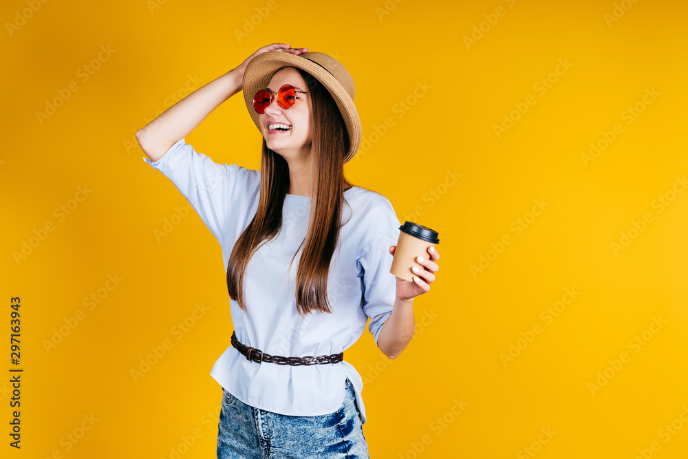 Pretty woman laughs with happiness in the studio with a yellow background. Dressed in a straw hat, jeans and a blouse. Holding a cup of coffee in her hand Pretty woman laughs with happiness in the studio with a yellow background. Dressed in a straw hat, jeans and a blouse. Holding a cup of coffee in her hand