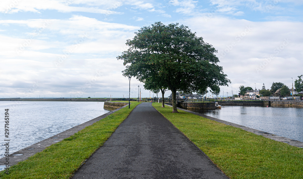 Fototapeta premium Footpath through park at Galway quay in the city, with trees, grass & sea on a sunny summer day.