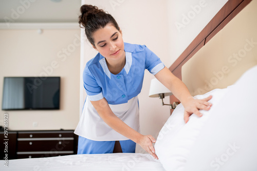 Pretty young professional chamber maid putting white clean pillows on bed