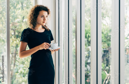 Young caucasian casual business woman standing thinking while drinking coffee beside home office window