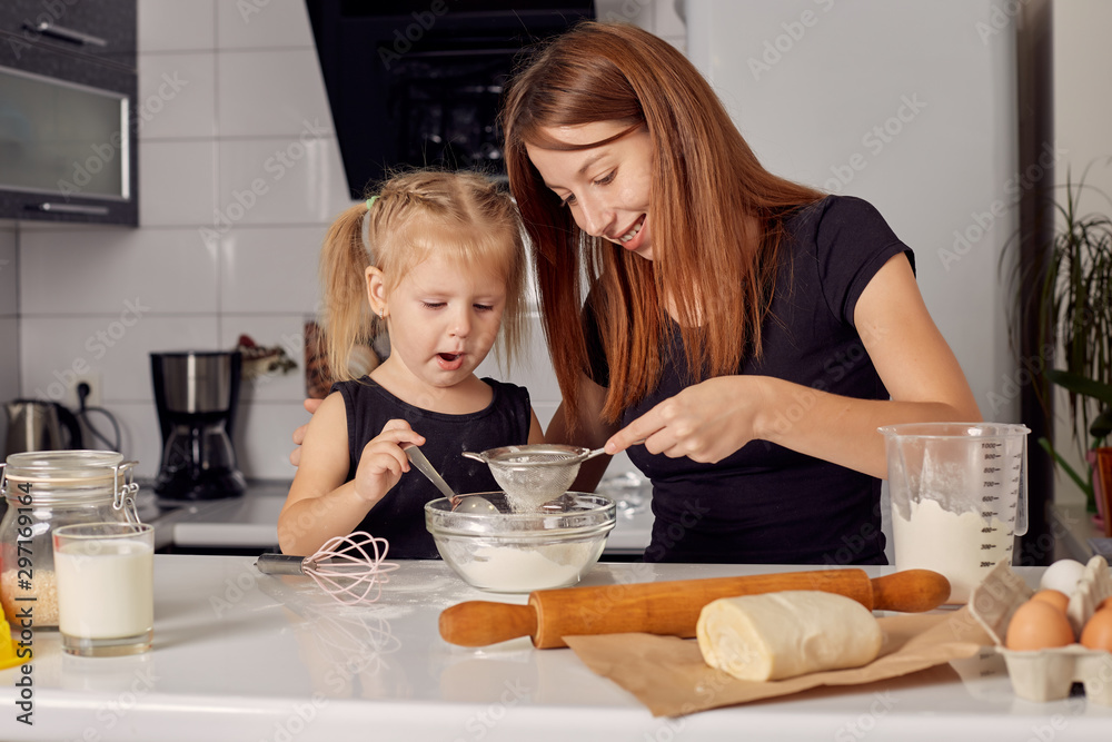 Mother and daughter preparing a sweet cake using flour, milk, sitting on chairs at a table in a modern kitchen. Girl holding a whisk, stirring eggs in a bowl, preparing pancake dough with her mom. Mother and daughter preparing a sweet cake using flour, milk, sitting on chairs at a table in a modern kitchen. Girl holding a whisk, stirring eggs in a bowl, preparing pancake dough with her mom.
