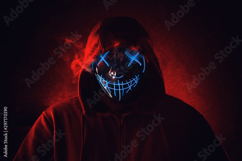 Man in angry and scary lighting neon glow mask in hood on dark red background. Halloween and horror concept