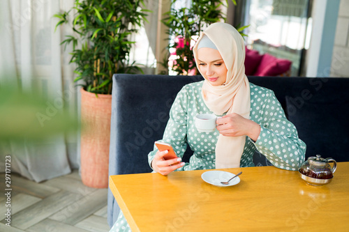 beautiful young woman in muslim dress is drinking tea in a restaurant with a phone in her hand