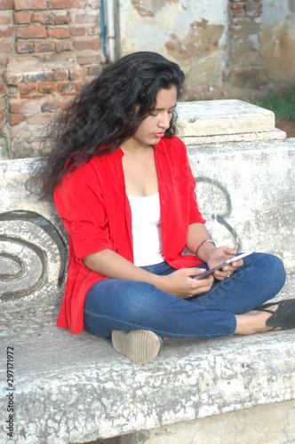Young wavy hair sitting on a semento bench with its feet between loops, looking straight at her tablet while smiling, wearing a red shirt over a white shirt and blue trousers. photo