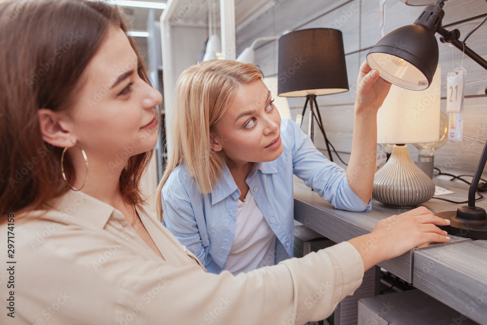 Obraz premium Two young women choosing new lamp to buy at furnishings store. Beauitful woman shopping for home lighting with her friend